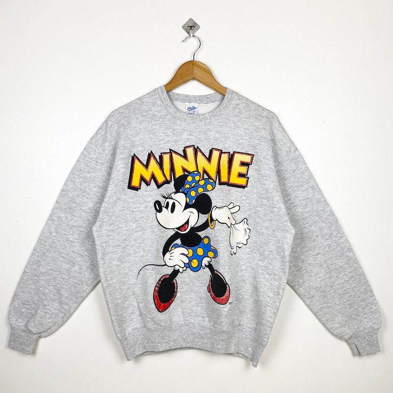 K&ouml;nnte beinhalten: Grauer Pullover mit einem Cartoon-Bild von Minnie Mouse in einem blau-gelb gepunkteten Kleid. Der Text "MINNIE" ist in Gelb &uuml;ber ihr gedruckt.