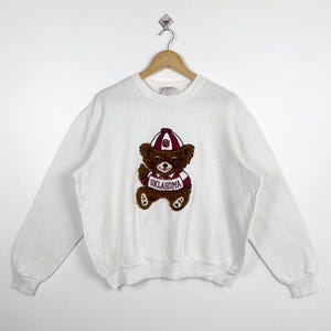 May include: White sweatshirt with a brown teddy bear wearing a red and white Oklahoma Sooners jersey and a red hat with a white "OU" logo.