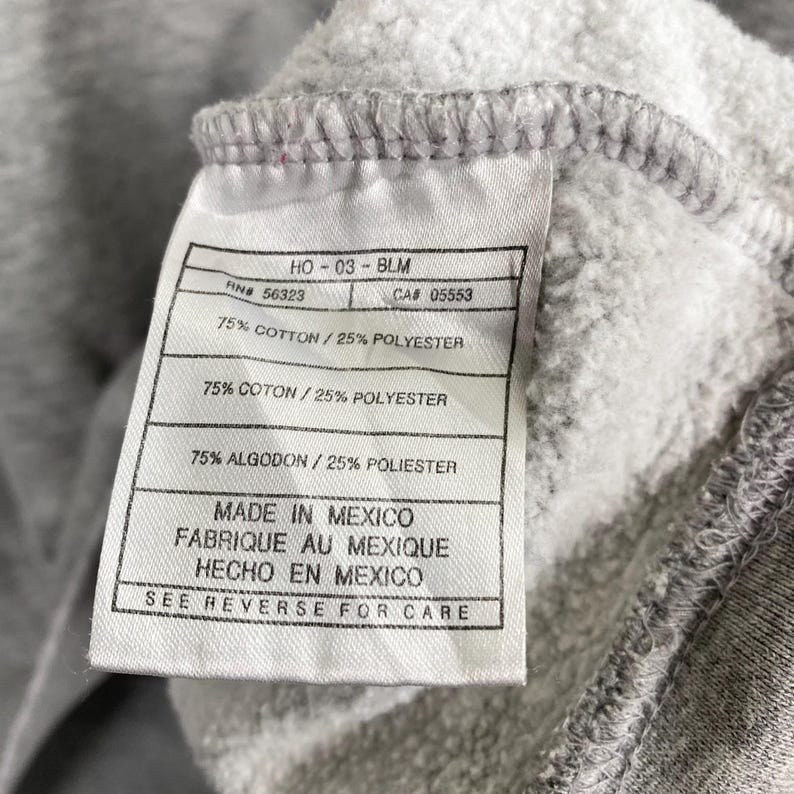 May include: Close-up of a gray fabric tag with black text. The tag reads "HO-03-BLM", "RN# 56323", "CA# 05553", and "75% COTTON / 25% POLYESTER". The tag also states "MADE IN MEXICO" and "SEE REVERSE FOR CARE".