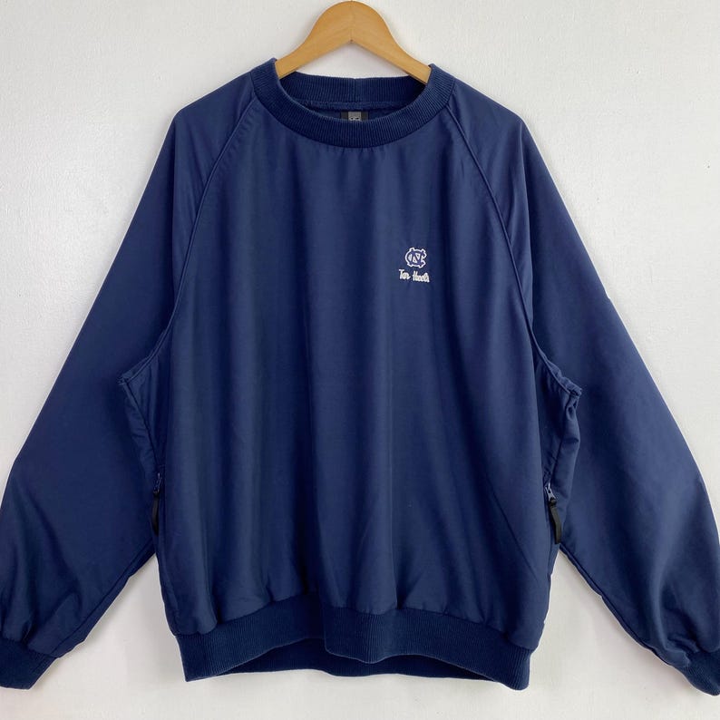 May include: Navy blue pullover jacket with a crew neck and long sleeves. Features a small white logo on the chest and side pockets with zippers. The jacket is hanging on a wooden hanger.