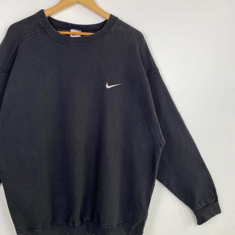 Puede incluir: Una sudadera Nike negra con un logotipo Swoosh blanco en el pecho izquierdo. La sudadera tiene cuello redondo y mangas largas.