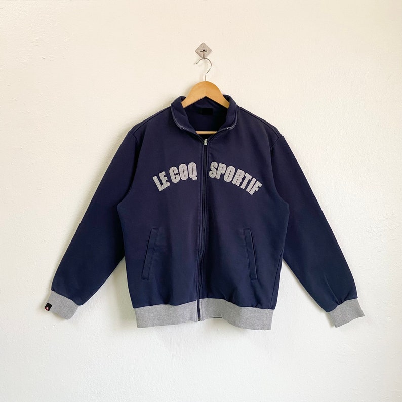 May include: A navy blue zip-up jacket with a gray ribbed hem and cuffs. The jacket has a white embroidered logo that reads "Le Coq Sportif".