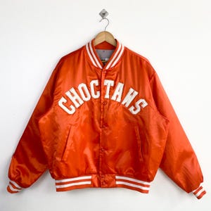 Op de afbeelding: Fel oranje satijnen jas met het woord "CHOCTAWS" in witte blokletters op de borst. De jas heeft wit en grijs gestreepte details op de kraag, manchetten en taille. Het is dichtgeknoopt en hangt aan een houten hanger.