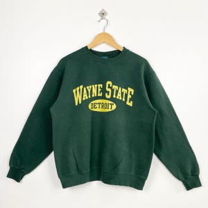 Może przedstawiać: Ciemnozielona bluza z napisem "WAYNE STATE DETROIT" w żółtym kolorze. Bluza wisi na wieszaku na białym tle. Tekst jest wygięty w łuk na piersi.