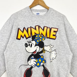 K&ouml;nnte beinhalten: Grauer Pullover mit einem Cartoon-Bild von Minnie Mouse in einem gelben gepunkteten Rock und einer gelben Schleife. Der Text "MINNIE" ist in gelben Buchstaben &uuml;ber ihr gedruckt.