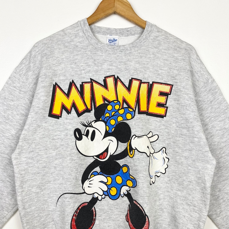 K&ouml;nnte beinhalten: Grauer Pullover mit einem Cartoon-Bild von Minnie Mouse in einem gelben gepunkteten Rock und einer gelben Schleife. Der Text "MINNIE" ist in gelben Buchstaben &uuml;ber ihr gedruckt.
