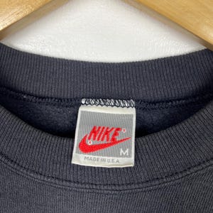 May include: A black Nike sweatshirt with a white tag that says "NIKE" in red with a white checkmark and "MADE IN U.S.A." in black. The tag also has a size "M" in black.