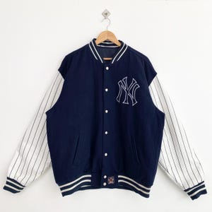 Può includere: Una giacca da baseball a righe blu navy e bianche con un grande logo "NY" ricamato sul davanti. La giacca ha una chiusura a zip e un colletto, polsini e orlo a coste.