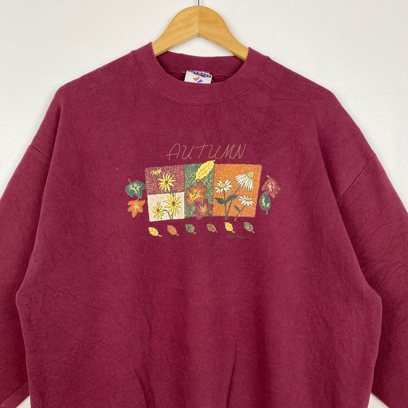 May include: A maroon sweatshirt with a graphic of autumn leaves and flowers. The text "AUTUMN" is printed above the graphic.