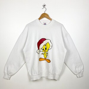 May include: White sweatshirt with a yellow Tweety Bird wearing a red Santa hat. The sweatshirt has a crew neck and long sleeves.