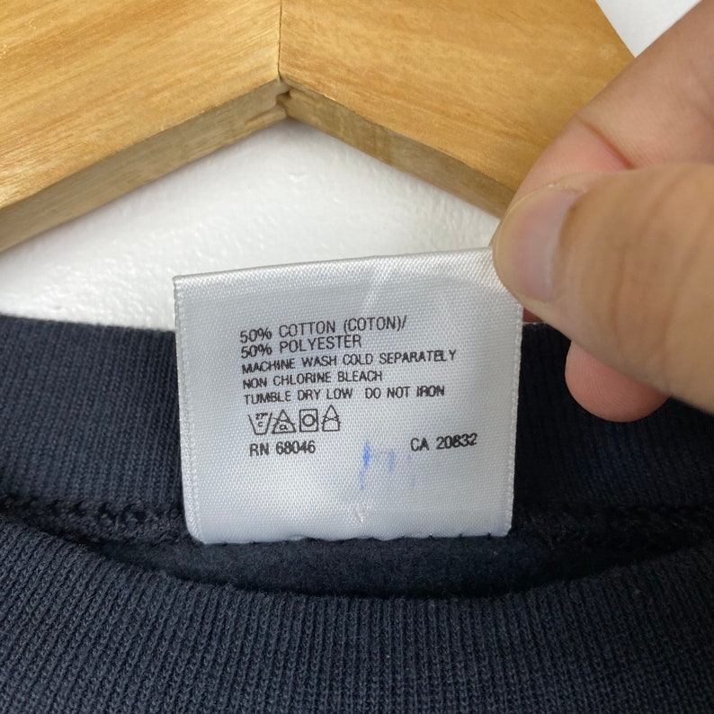 May include: White care label with black text detailing the fabric content and care instructions for a garment. The label reads: 50% Cotton (Coton)/ 50% Polyester Machine Wash Cold Separately Non Chlorine Bleach Tumble Dry Low Do Not Iron RN 68046 CA 20832