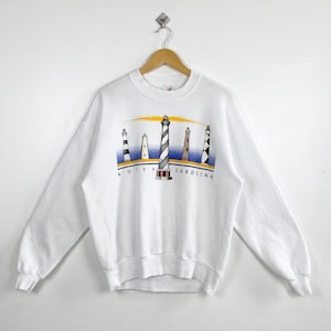 May include: White sweatshirt featuring a colorful graphic of five lighthouses under a yellow and orange sky. The text "NORTH CAROLINA" is printed below the lighthouses. The sweatshirt has long sleeves and a crew neck.