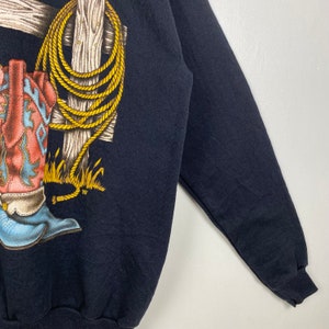May include: A black long-sleeved sweatshirt with a graphic print of a cowboy boot and a rope wrapped around a wooden post. The boot is red and blue with a yellow star on the side.