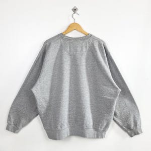 May include: A heather gray sweatshirt hangs on a wooden hanger against a white background. The crew neck sweatshirt has long sleeves and a ribbed hem and cuffs. The back of the sweatshirt is visible, showing the neckline and shoulder seams.