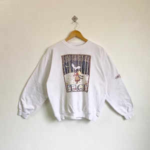 Puede incluir: Sudadera blanca de manga larga con un diseño que representa una escena de invierno con una niña sosteniendo un pájaro y la palabra "PEACE" impresa debajo. La sudadera tiene un pequeño diseño bordado en la manga.