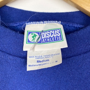 May include: A blue sweatshirt with a white tag that reads "DISCUS ATHLETIC" and "Medium" in black text. The tag also includes the text "MADE IN U.S.A. / HECHO EN E.U.A.J FABRIQUE AUX E.-U. Mediano/Moyen/サイズ:M".