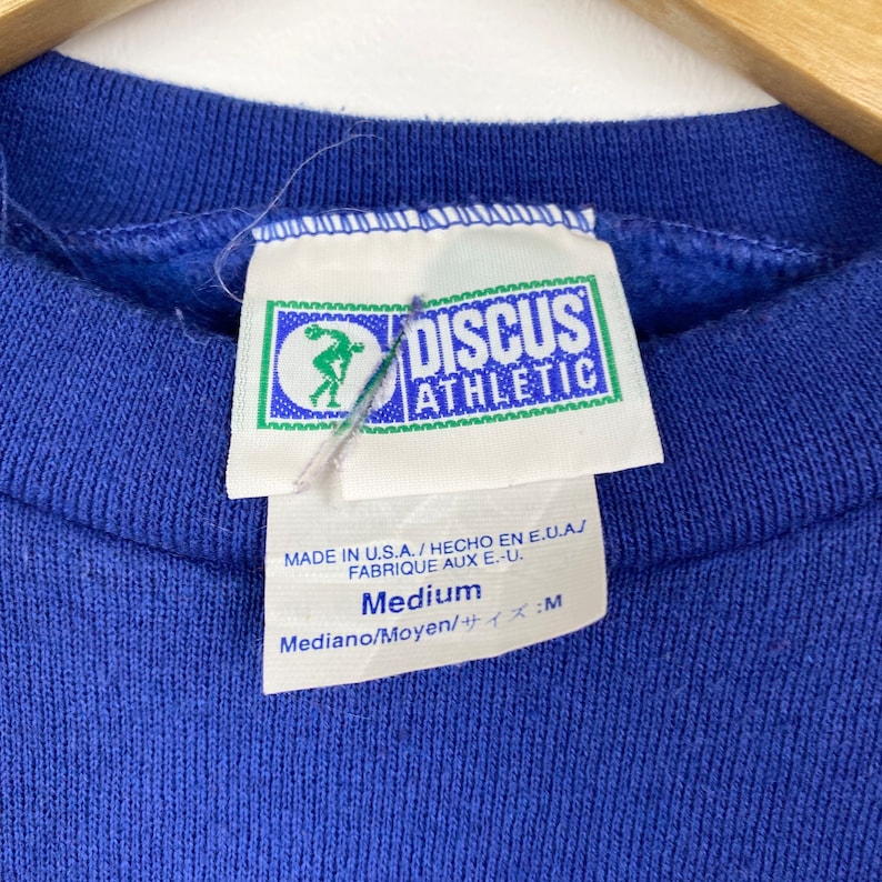 May include: A blue sweatshirt with a white tag that reads "DISCUS ATHLETIC" and "Medium" in black text. The tag also includes the text "MADE IN U.S.A. / HECHO EN E.U.A.J FABRIQUE AUX E.-U. Mediano/Moyen/サイズ:M".