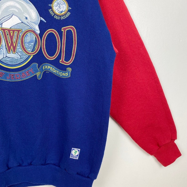May include: Blue and red sweatshirt with a dolphin and the text "WOOD" in a circle with the text "EXPEDITIONS" and "NEW JERSEY" below. The text "SAVE OUR OCEANS" is above the circle. The sweatshirt has a green label with the text "INSCUS".