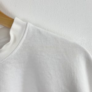 May include: Close-up of a white crew neck sweatshirt. The ribbed fabric is visible, and the neckline is clean and simple. The sweatshirt is hanging on a wooden hanger against a white wall.