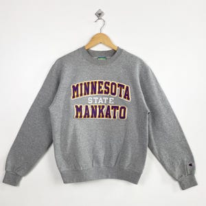 Op de afbeelding: Een grijze sweatshirt met de tekst "MINNESOTA STATE MANKATO" in goud en paars. De sweatshirt hangt aan een houten hanger tegen een witte achtergrond. Het heeft lange mouwen en een ronde hals.