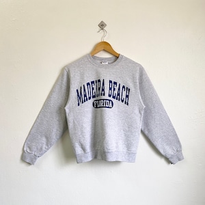 May include: A gray crewneck sweatshirt with the text "MADEIRA BEACH FLORIDA" in navy blue arched across the chest. The sweatshirt is hanging on a wooden hanger against a white wall.