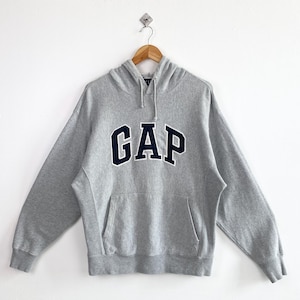 Könnte beinhalten: Grauer Kapuzenpullover mit dem Wort "GAP" in marineblauen Großbuchstaben auf der Brust. Der Pullover hat eine Vordertasche und eine Kapuze mit Kordelzug. Er hängt an einem Holzbügel vor weißem Hintergrund.