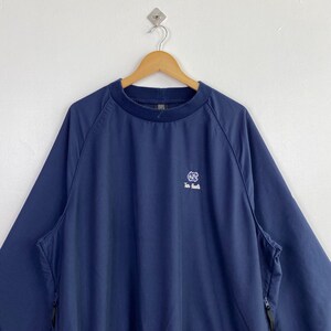 May include: Navy blue pullover jacket with a crew neck and long sleeves. The jacket features a small white logo on the chest and two side pockets with zippers. The jacket is hanging on a wooden hanger against a white background.