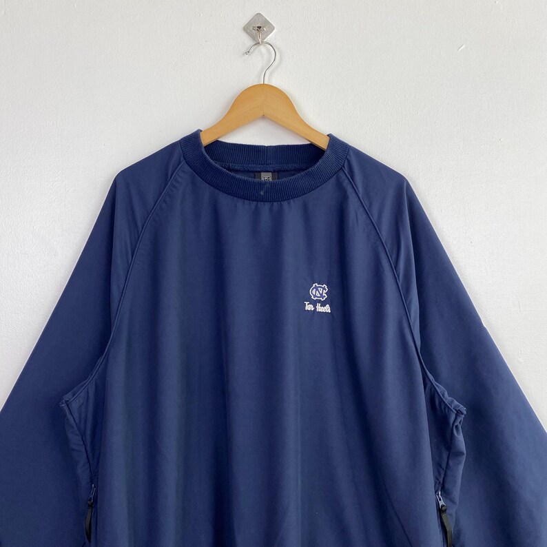 May include: Navy blue pullover jacket with a crew neck and long sleeves. The jacket features a small white logo on the chest and two side pockets with zippers. The jacket is hanging on a wooden hanger against a white background.