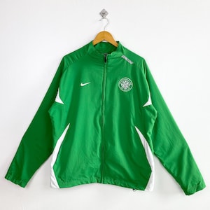May include: Green and white athletic jacket with a full zipper. The jacket features a white Nike logo on the chest and white accents on the sleeves. A circular emblem is on the right chest. The jacket is hanging on a wooden hanger.