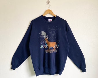 May include: A navy blue sweatshirt with a graphic print of a deer standing in front of a full moon. The deer is brown with white spots and has large antlers. The graphic also includes a stylized design of a Native American rug.