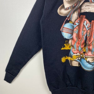 May include: Black long-sleeve sweatshirt with a graphic print of cowboy boots, a hat, and a canteen.