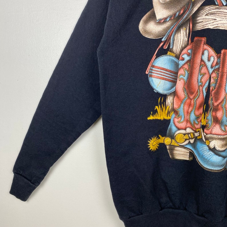May include: Black long-sleeve sweatshirt with a graphic print of cowboy boots, a hat, and a canteen.