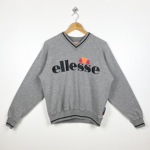 May include: A gray v-neck sweatshirt with a black ellesse logo and a red, orange, and white logo. The sweatshirt has black trim around the neck and cuffs.