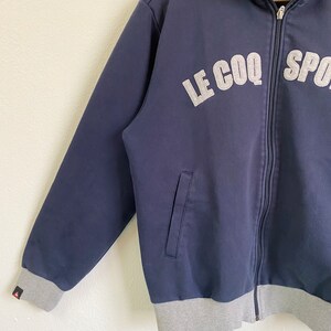 May include: A navy blue zippered jacket with a gray ribbed hem and cuffs. The jacket has a silver embroidered logo that reads "LE COQ SPORT".