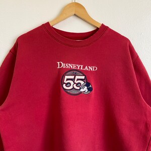 May include: Red sweatshirt with a white Disneyland logo and a Mickey Mouse graphic with the text "Est. 55".