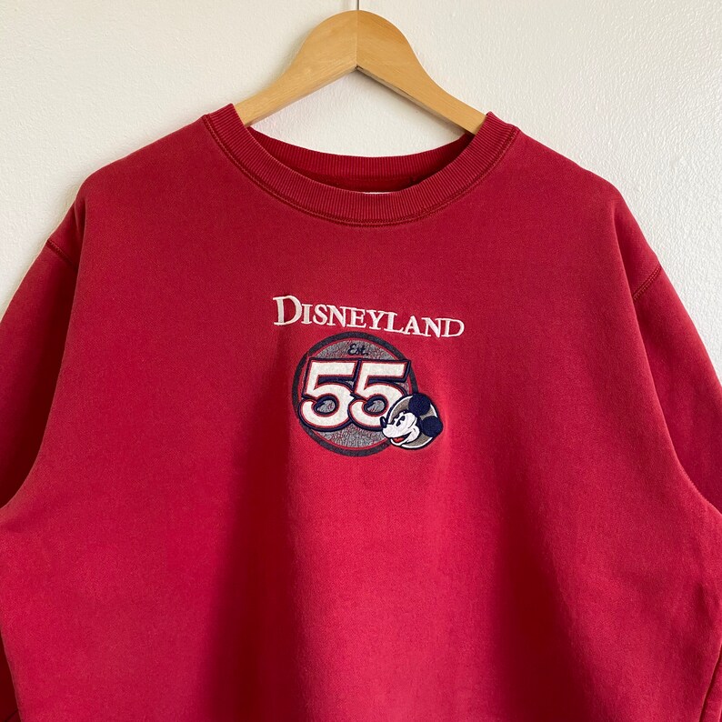 May include: Red sweatshirt with a white Disneyland logo and a Mickey Mouse graphic with the text "Est. 55".