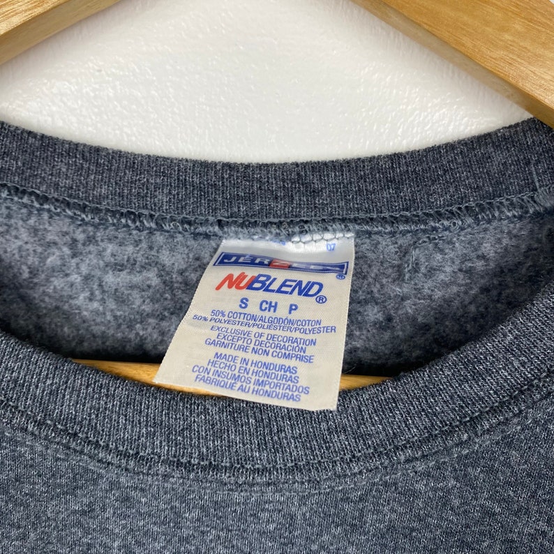 May include: A grey sweatshirt tag with the text "NUBLEND" in a white box with a black border. The tag also includes the text "S CH P" and the following information: "50% Cotton/Algod&oacute;n/Cotton 50% Polyester/Poli&eacute;ster/Polyester Exclusive of Decoration Excepto Decoraci&oacute;n Garniture Non Comprise Made in Honduras Hecho en Honduras Con Insumos Importados Fabrique au Honduras"