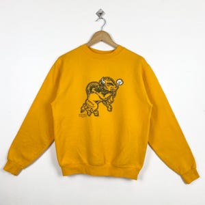 May include: Mustard yellow sweatshirt featuring a graphic of a bison with the text "NDSU" and "NORTH DAKOTA STATE". The sweatshirt has a crew neck and long sleeves. The graphic is in shades of brown and tan.