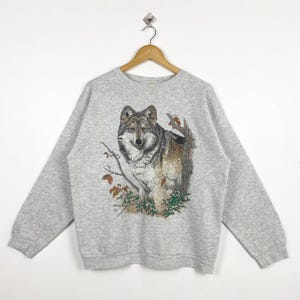May include: A light grey sweatshirt with a detailed wolf graphic. The wolf is shown with realistic fur, set against trees and foliage. The sweatshirt has long sleeves and a crew neck. The design is printed on the front.