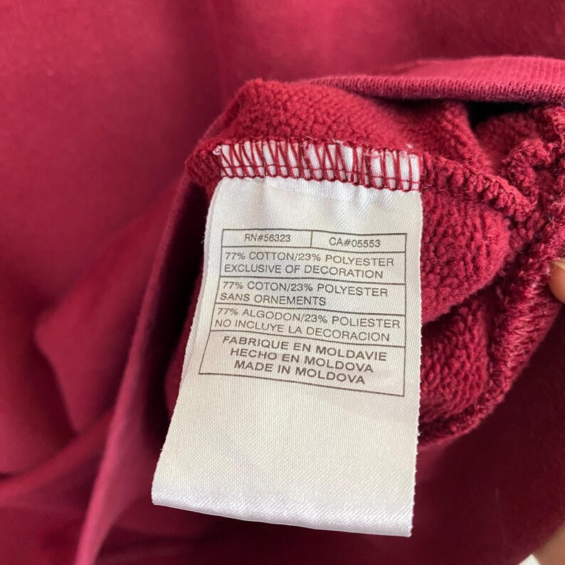 May include: A white tag with black text on a maroon sweatshirt. The tag lists the fabric content as 77% cotton and 23% polyester. The tag also states that the decoration is exclusive of the fabric content. The tag includes the RN number 56323 and the CA number 05553. The tag also states that the garment was made in Moldova.