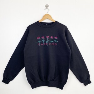 Puede incluir: Sudadera negra con bordado de rosas rosas y el texto "CAPE COD" en turquesa. La sudadera de cuello redondo y manga larga tiene un corte relajado. El diseño está centrado en el pecho.