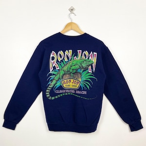 Op de afbeelding: Een donkerblauw sweatshirt met lange mouwen met een grafische print van een groene leguaan op een tak. De grafiek bevat de tekst "Ron Jon Surf Shop Clearwater Beach".