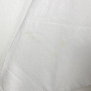 May include: Close-up of a white fabric with a slight sheen. The fabric has a few small, light-brown stains. The material appears to be smooth and possibly a lightweight textile, such as cotton or linen.