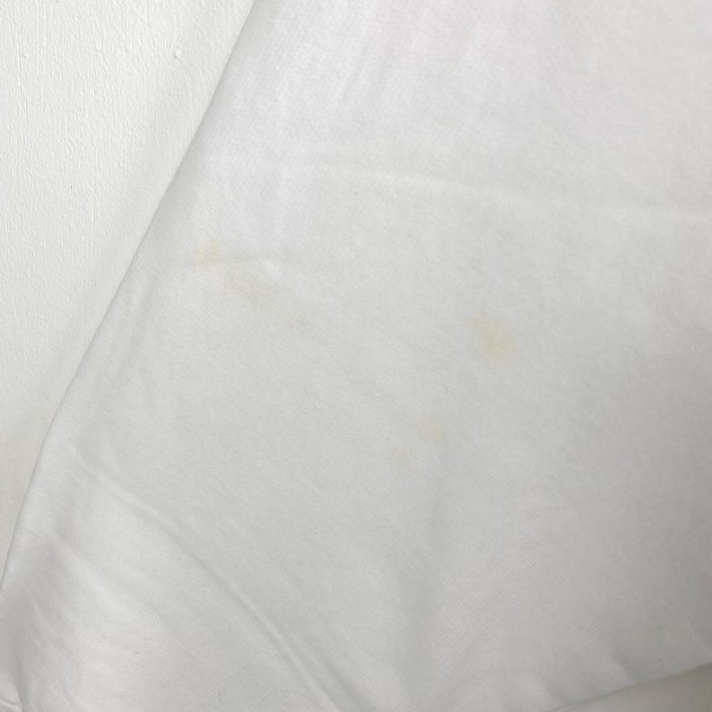 May include: Close-up of a white fabric with a slight sheen. The fabric has a few small, light-brown stains. The material appears to be smooth and possibly a lightweight textile, such as cotton or linen.
