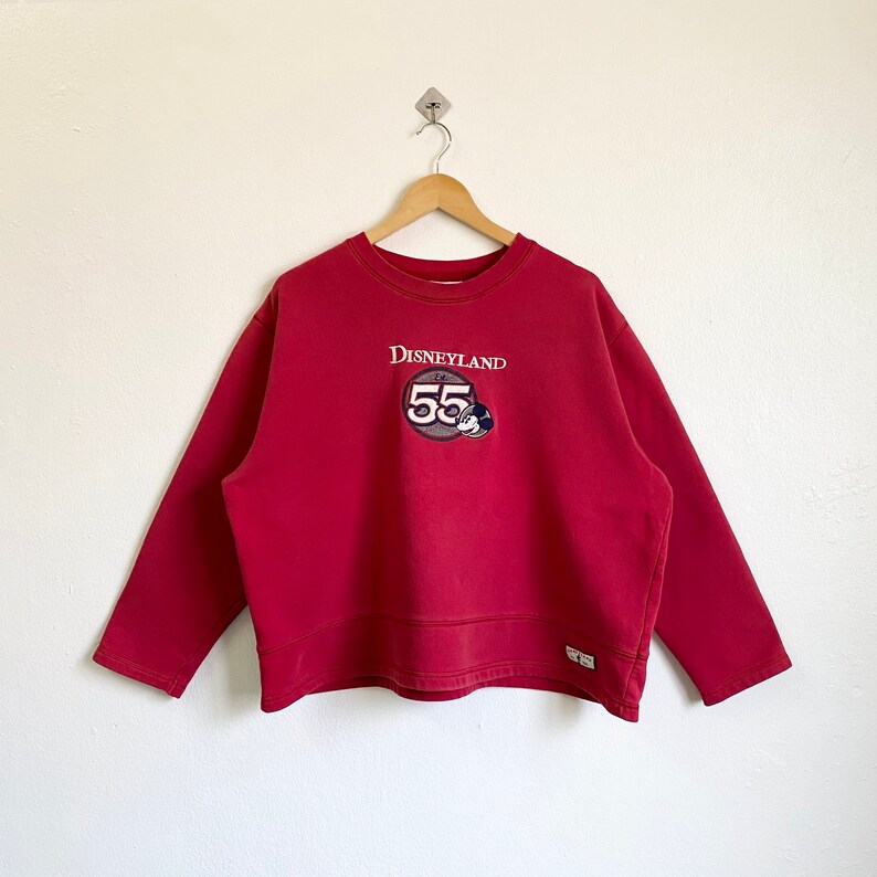 May include: Red long-sleeve sweatshirt with a vintage Disneyland 55th anniversary logo featuring Mickey Mouse. The logo is a circular patch with a white border and the text "Disneyland" in white.