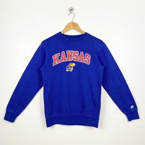 May include: A blue sweatshirt with the word "KANSAS" in red and white lettering and a red and yellow mascot in the centre. The sweatshirt has a white "R" logo on the right sleeve.