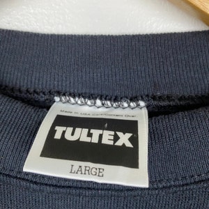 May include: A white tag with black text that reads "Made in USA Care/Content Over. Tultex Large" on a dark gray sweatshirt.