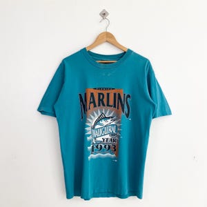 May include: A teal blue t-shirt with a graphic print of a marlin fish and the text "Florida Marlins Inaugural Year 1993".