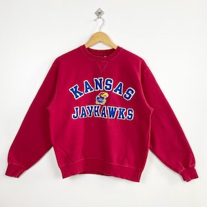 Puede incluir: Una sudadera roja de cuello redondo con el texto "KANSAS JAYHAWKS" en letras azules y blancas. La sudadera presenta un pequeño gráfico de la mascota Jayhawk. La prenda está colgada en una percha de madera sobre un fondo blanco.