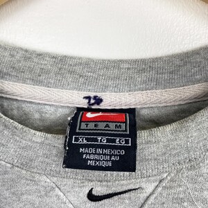 May include: Close-up of a gray crew neck sweatshirt with a black Nike swoosh logo at the bottom. A navy blue tag with the word "TEAM" and the Nike logo is visible. The tag also reads "MADE IN MEXICO FABRIQUE AU MEXIQUE".